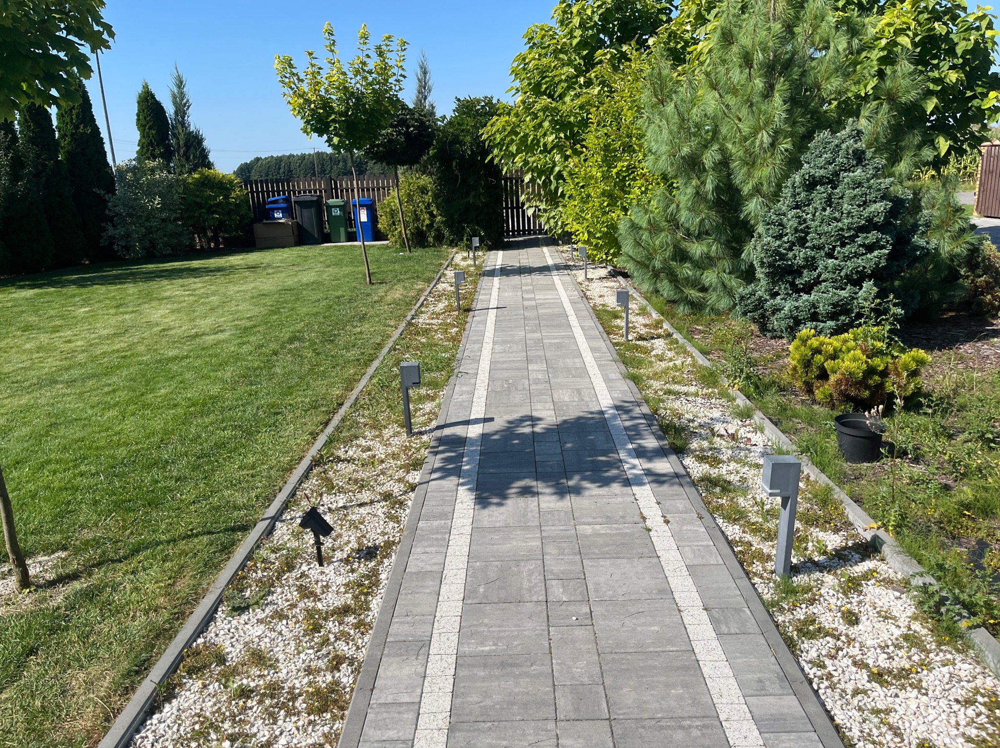 Ścieżka z kostki brukowej z oświetleniem w ogrodzie, otoczona trawnikiem i roślinnością. W tle widoczne pojemniki na śmieci i ogrodzenie.