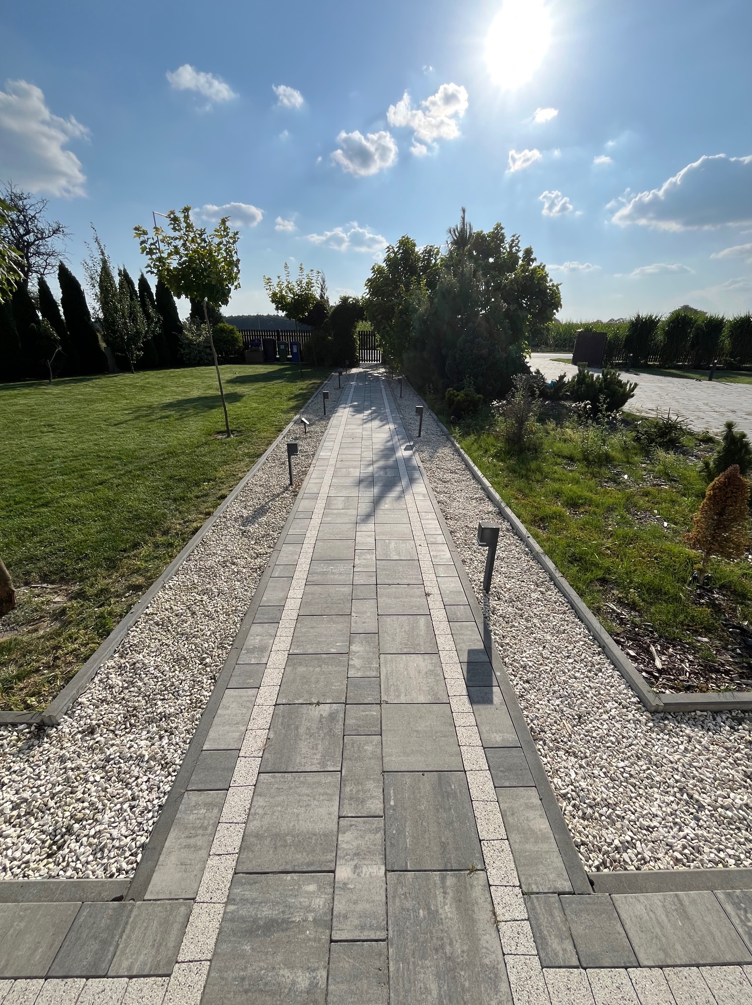 Ułożona kostka brukowa tworząca ścieżkę w ogrodzie z oświetleniem, otoczona białym żwirem i zielenią. Słoneczny dzień, błękitne niebo z chmurami.