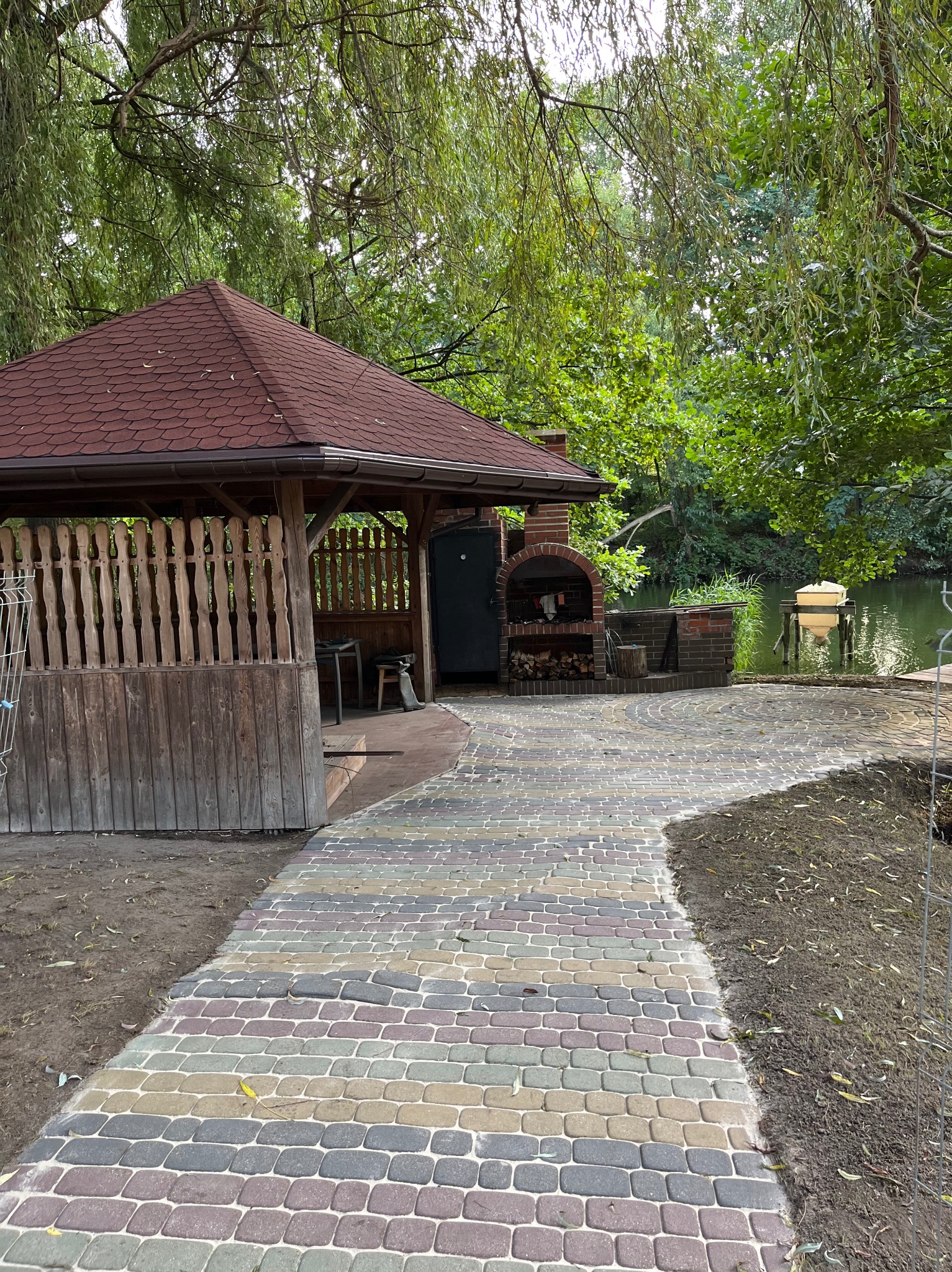 Kolorowa kostka brukowa prowadzi do drewnianej altany z grillem, usytuowanej nad brzegiem wody, otoczonej zielenią drzew.