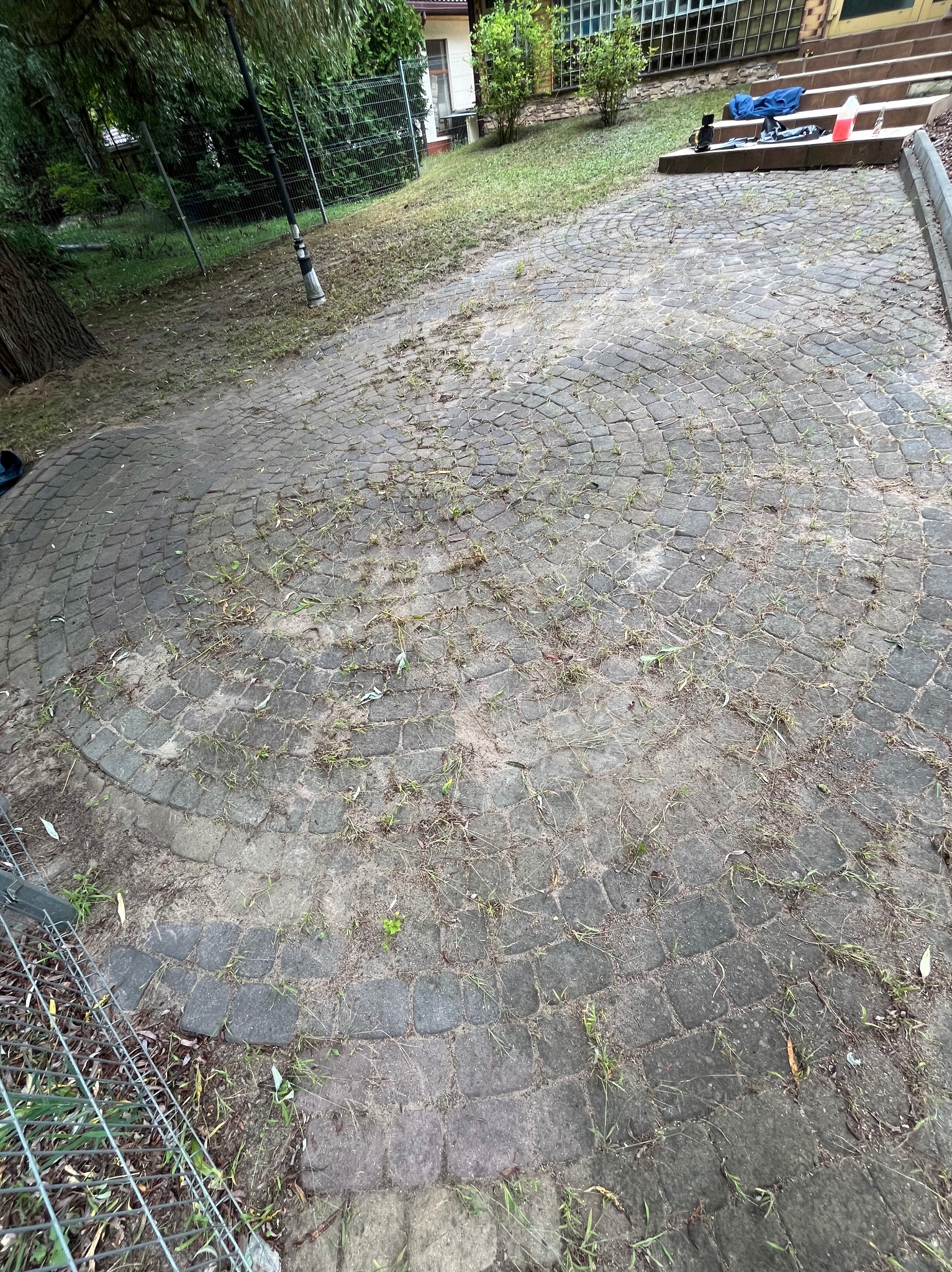 Zaniedbana, okrągła kostka brukowa z chwastami, wymagająca czyszczenia. Widoczne narzędzia i pojemnik, sugerujące przygotowania do pracy.