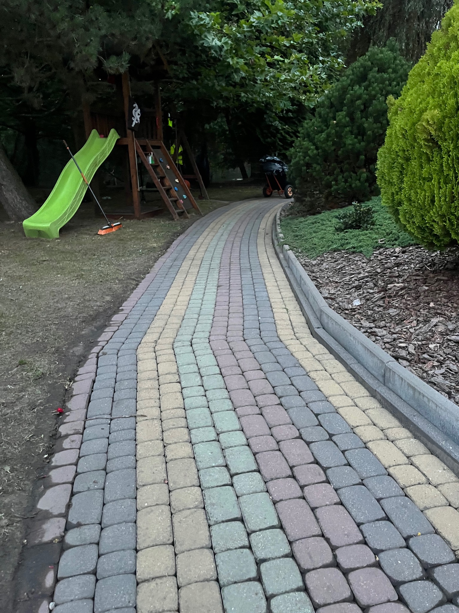 Kolorowa kostka brukowa tworząca ścieżkę w ogrodzie, prowadząca obok trawnika i zieleni, z placem zabaw w tle. Widoczne narzędzie do zamiatania.