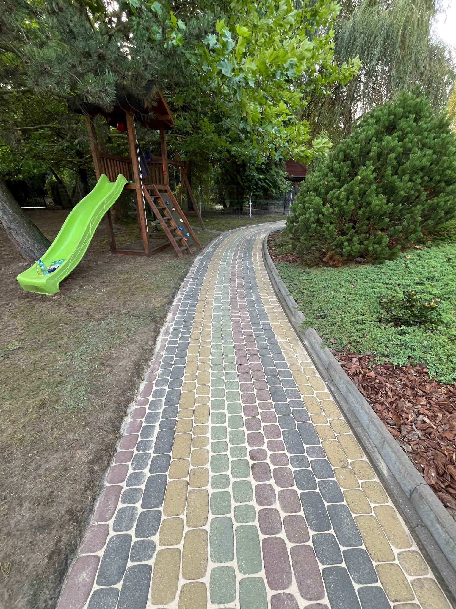 Kolorowa kostka brukowa tworzy krętą ścieżkę w ogrodzie z placem zabaw, zjeżdżalnią i bujną zielenią. Krawężnik oddziela ścieżkę od rabat z korą.