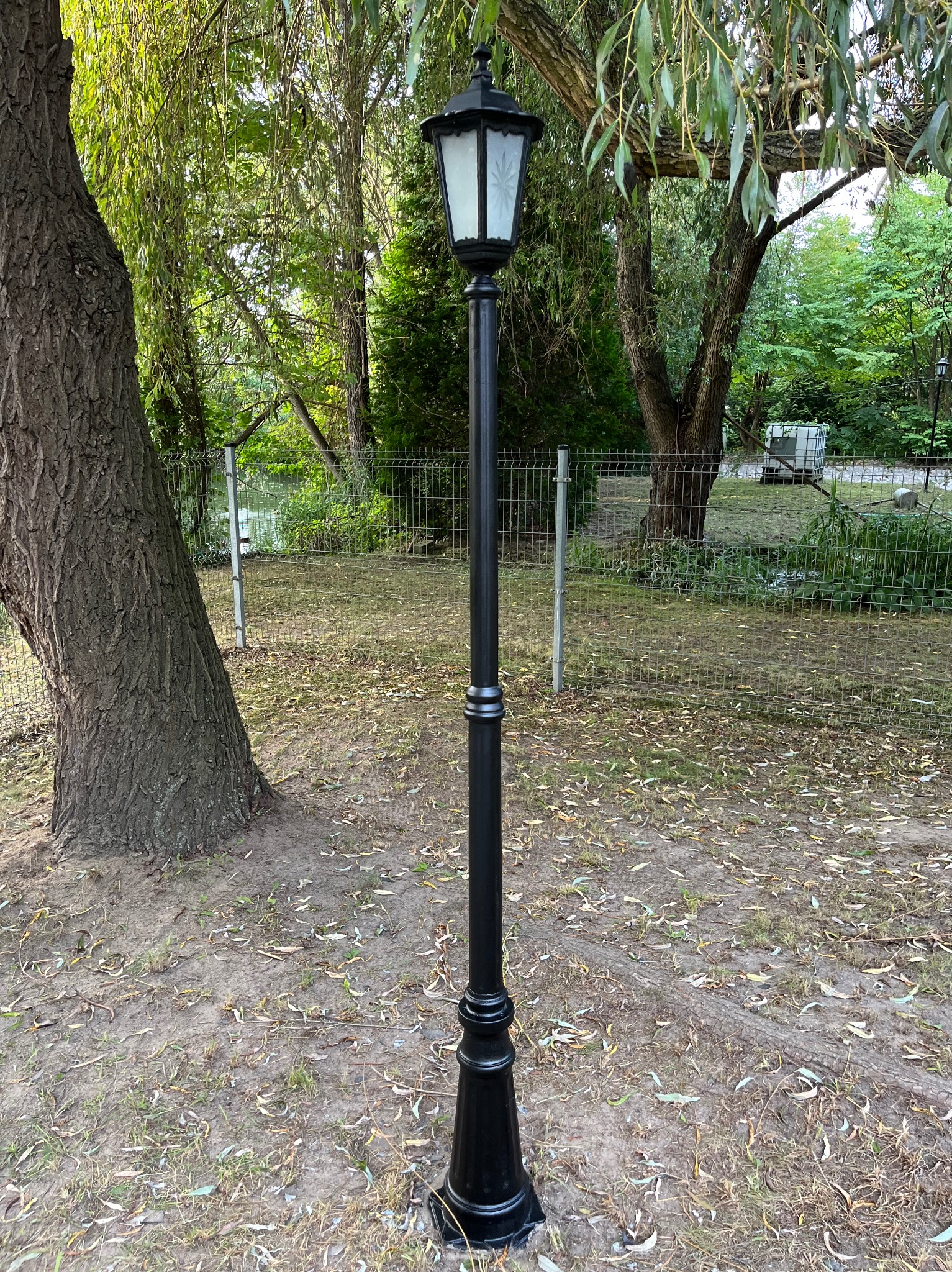 Czarna, wysoka latarnia parkowa z białym kloszem, umieszczona na trawiastym terenie z drzewami w tle. Metalowe ogrodzenie w oddali.
