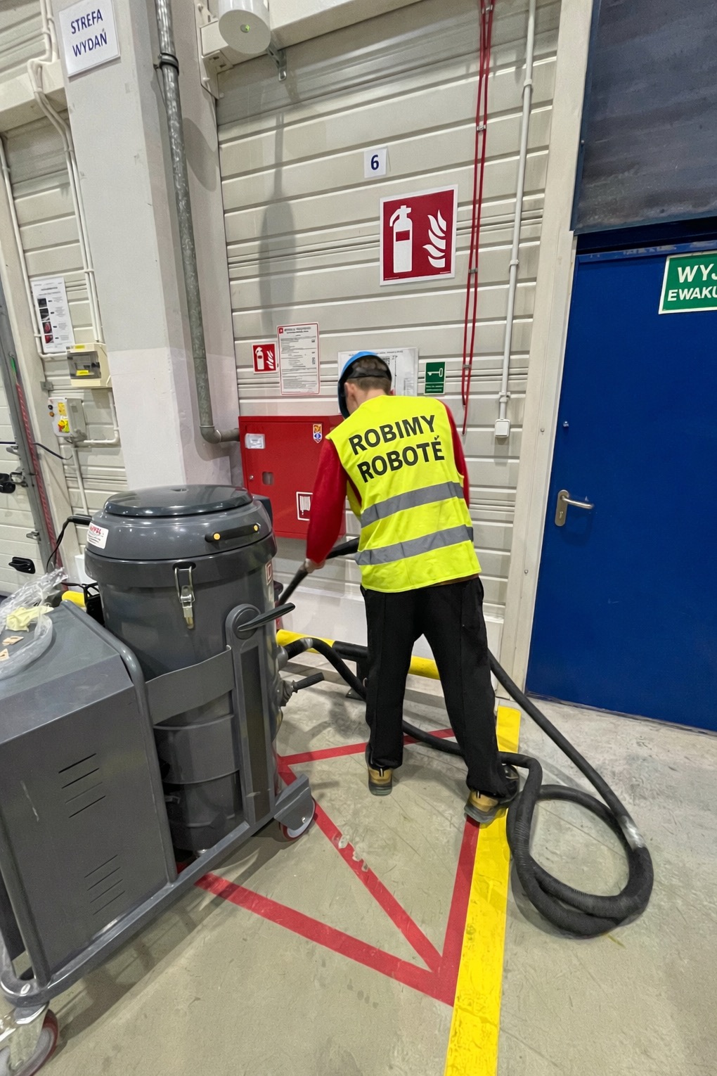 Pracownik w kamizelce z napisem 'ROBIMY ROBOTĘ' obsługuje przemysłowy odkurzacz w hali. Widoczny sprzęt ppoż. i oznakowania. Hala z betonową podłogą.