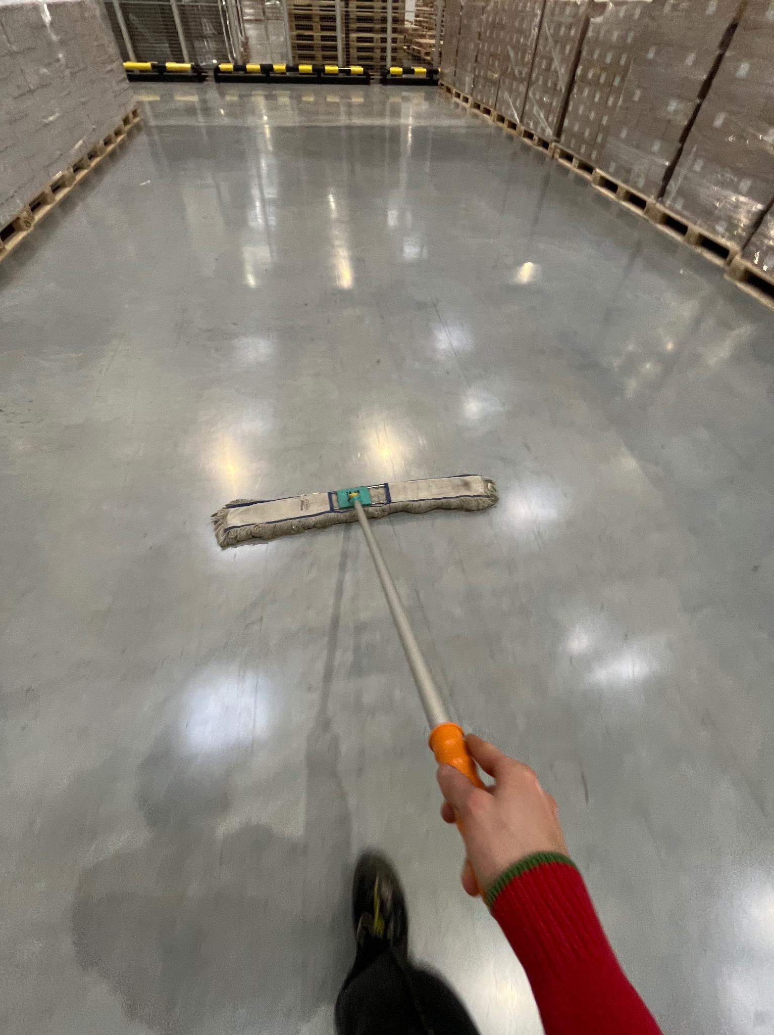 Mop na długim kiju czyszczący betonową posadzkę w magazynie. W tle palety z towarem. Widok z perspektywy osoby sprzątającej, ubrana w czerwoną bluzę.