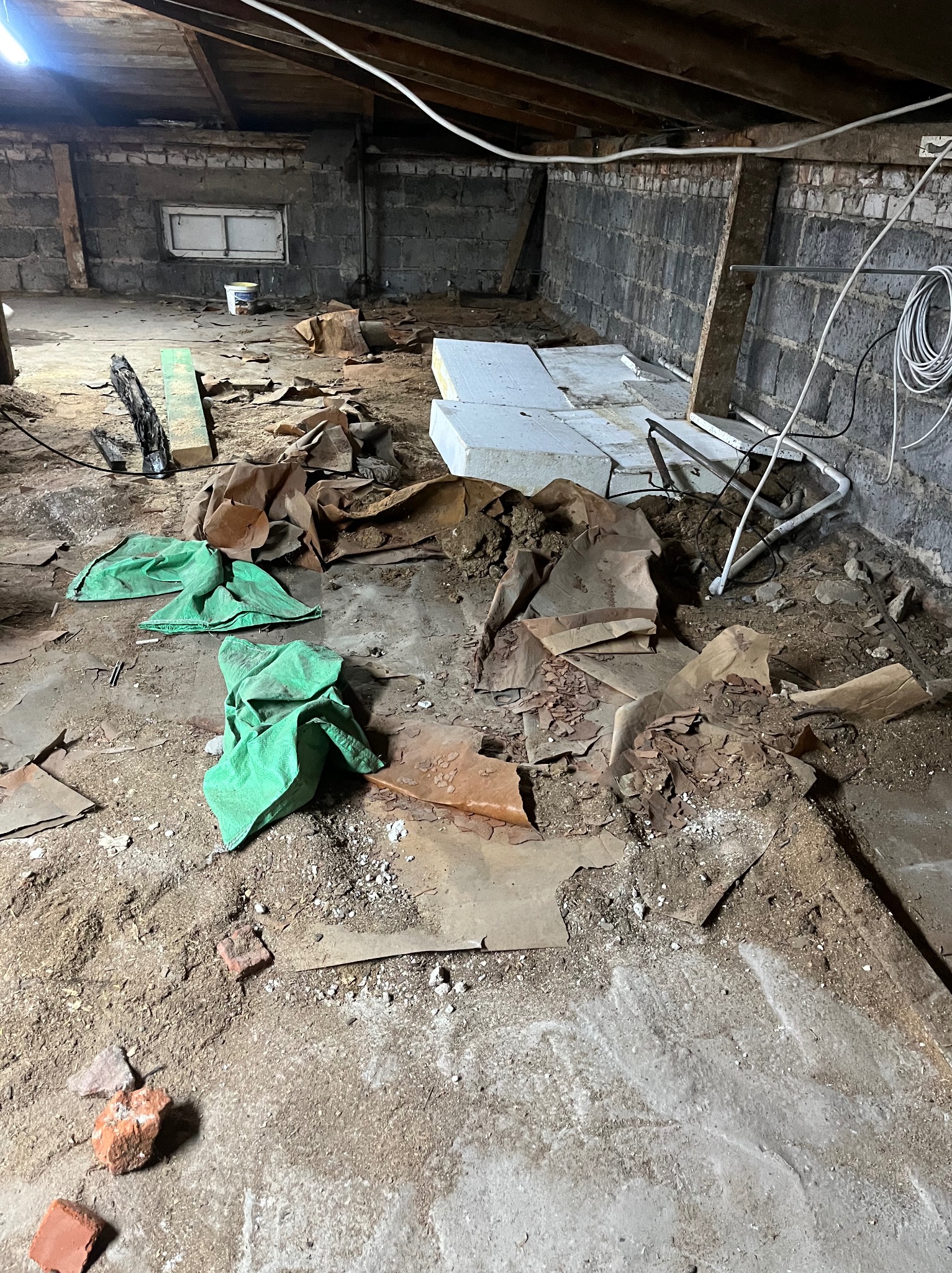 Piwnica w trakcie sprzątania po remoncie: gruz, folie, styropian, worki i resztki materiałów budowlanych na podłodze. Widoczne ślady po pracach remontowych na ścianach.