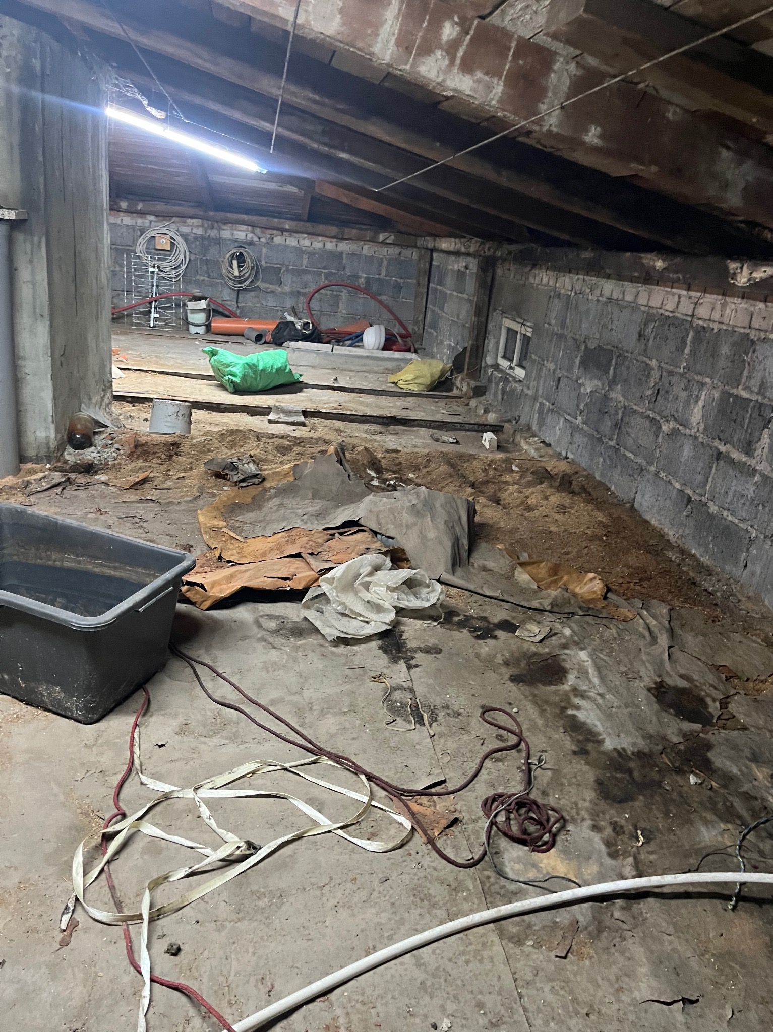 Zaniedbane poddasze z gruzem, kablami i workami. Widoczne ceglane ściany, drewniany strop i betonowy filar. W tle narzędzia i rury. Niezagospodarowana przestrzeń przed remontem.