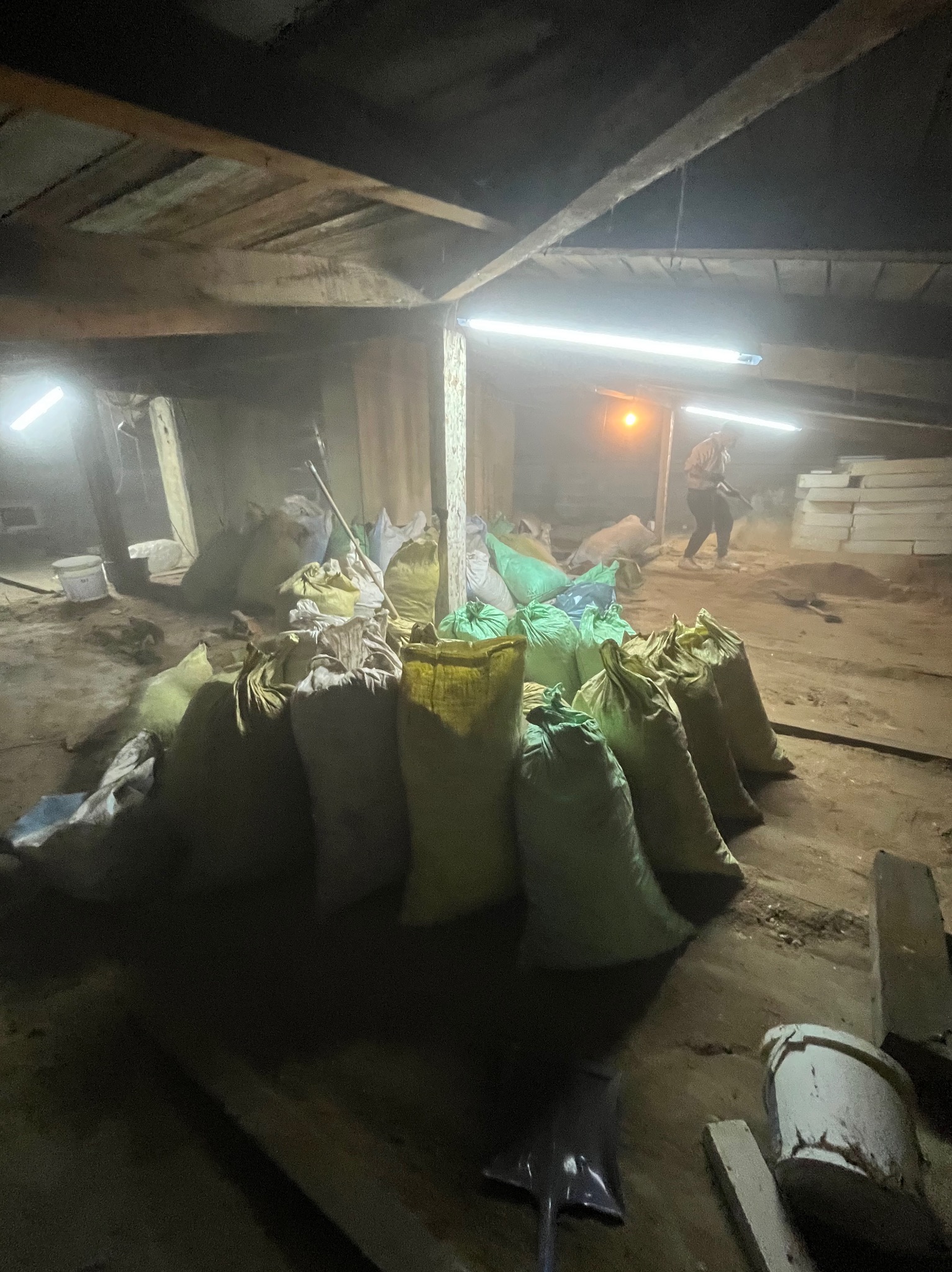 Wnętrze poddasza z workami gruzu i pracownikiem w tle, przygotowane do wywozu po remoncie. Widoczne narzędzia i prowizoryczne oświetlenie.