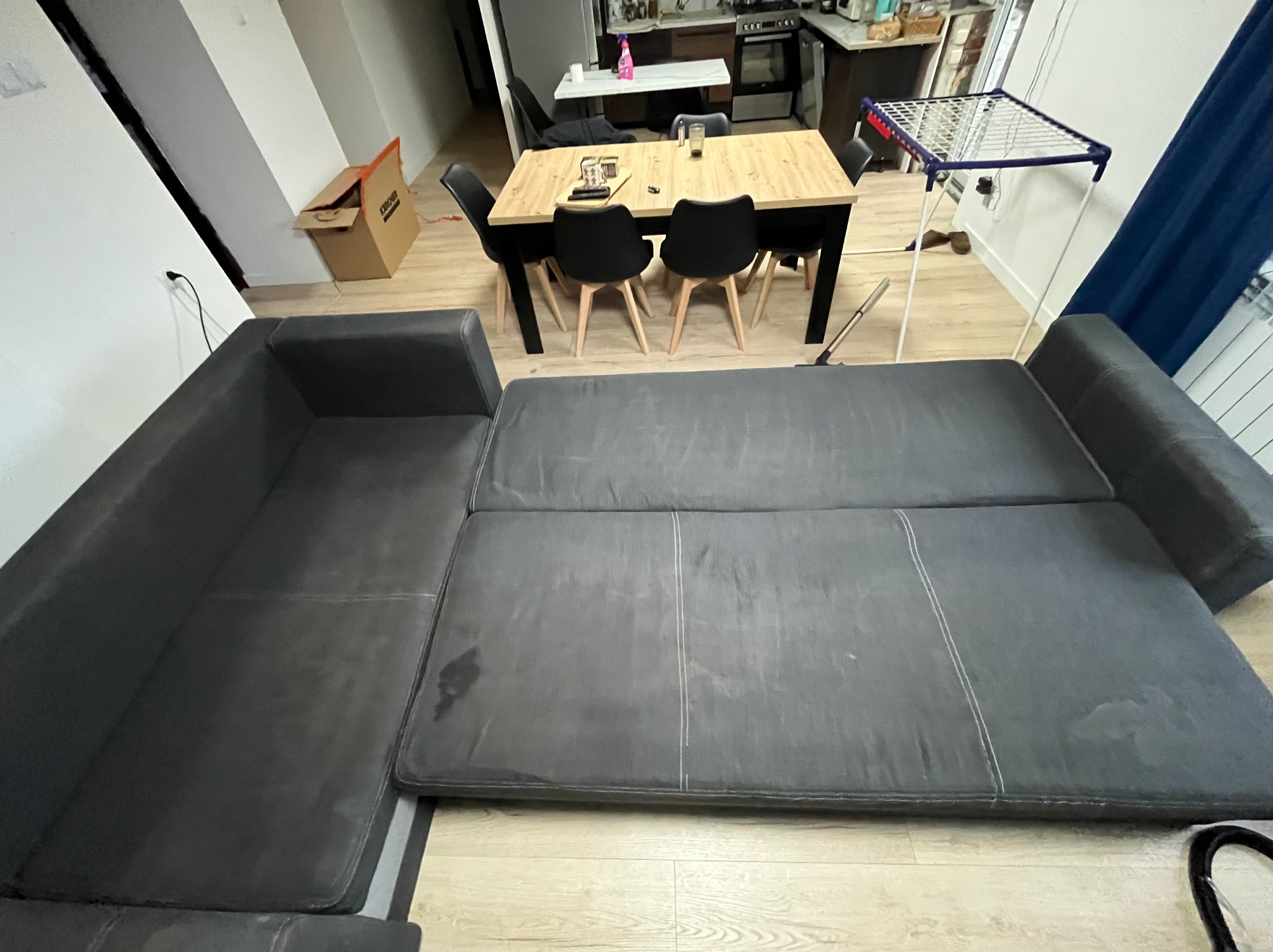 Szary narożnik z plamami na siedzisku w pokoju z drewnianym stołem i krzesłami. W tle widoczna kuchnia i suszarka na pranie. Narożnik wymaga czyszczenia.