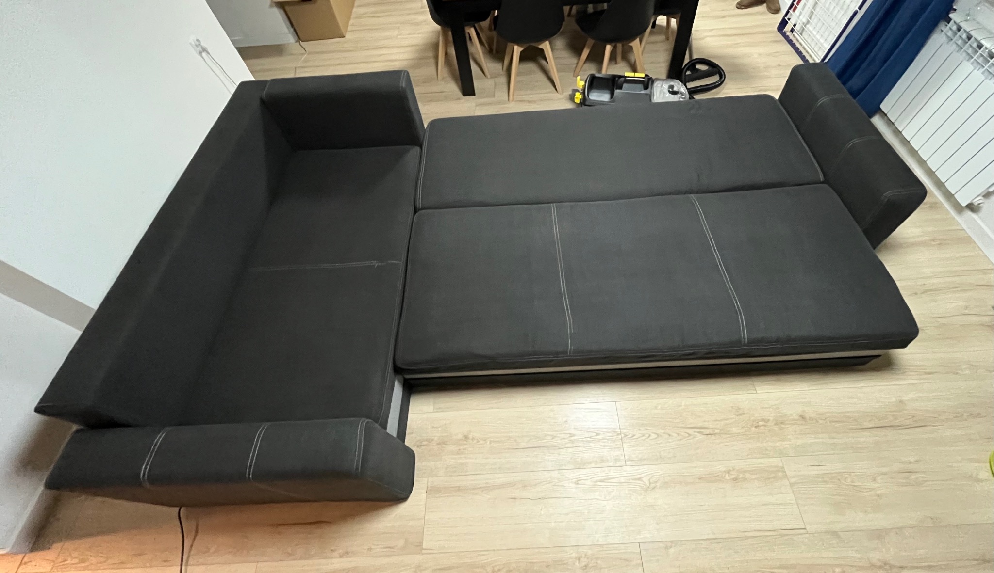 Rozłożony, grafitowy narożnik z widocznymi przeszyciami, ustawiony na panelach podłogowych. W tle stół z krzesłami i grzejnik.