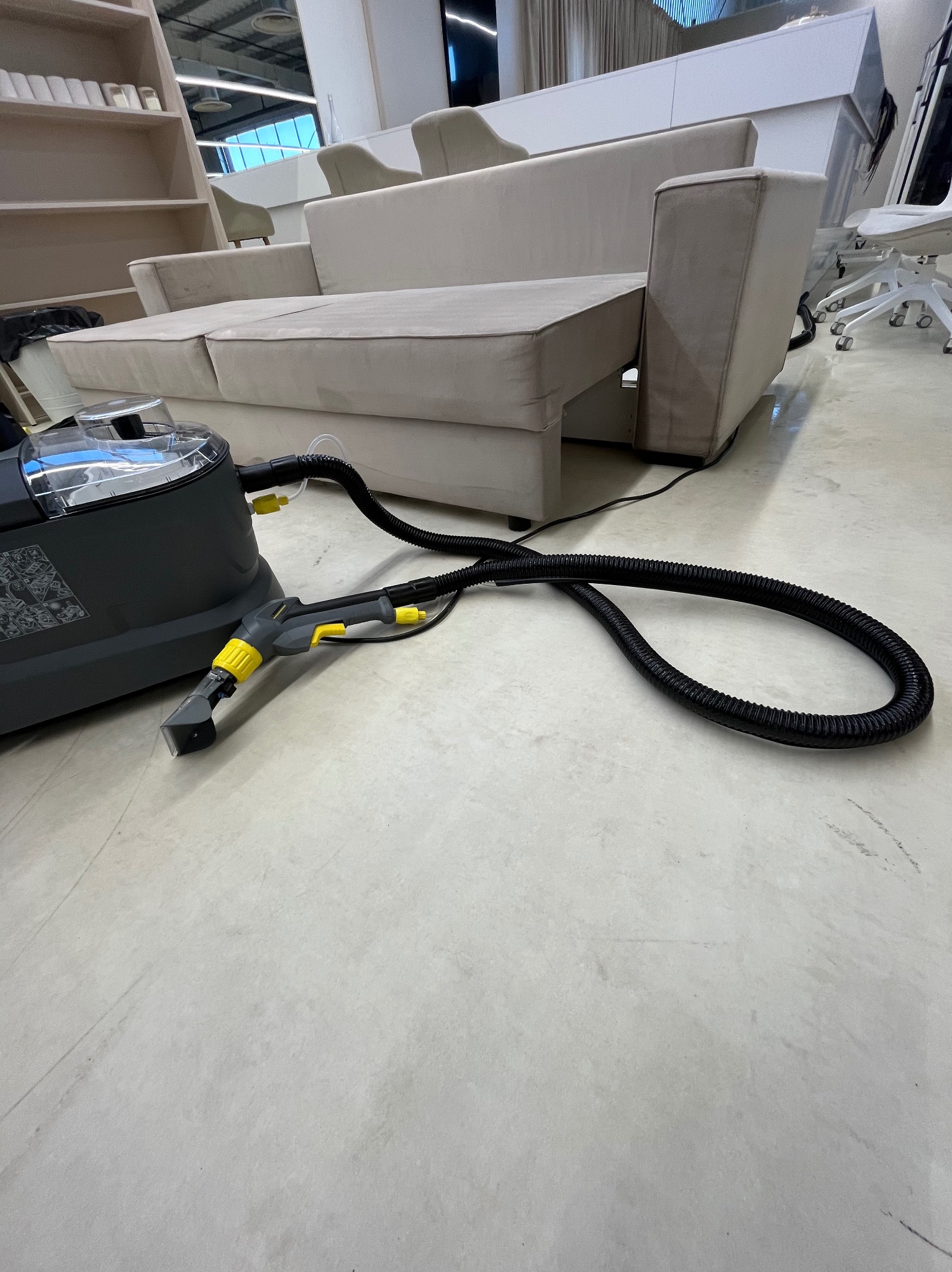 Profesjonalny odkurzacz piorący Karcher z czarnym wężem i dyszą na jasnej podłodze, obok beżowej sofy w nowoczesnym wnętrzu biurowym.