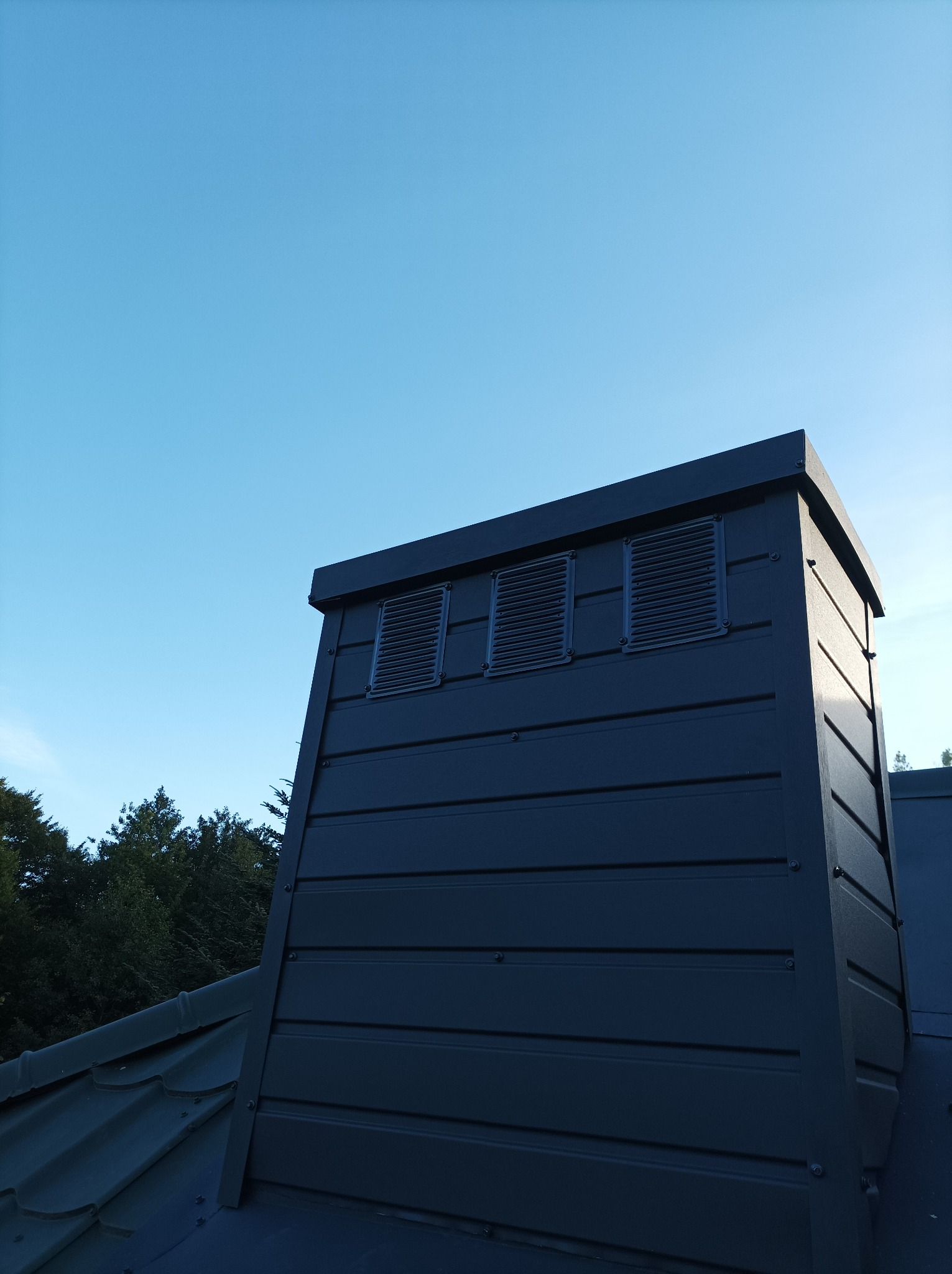 Nisko kątowe ujęcie grafitowego komina wentylacyjnego na dachu z blachy, z trzema kratkami wentylacyjnymi. W tle błękitne niebo i fragmenty zielonych drzew.