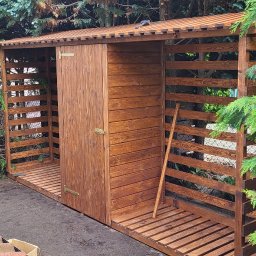 INSTAL-WOOD WIESŁAW SZWED - Drewniana, brązowa wiata ogrodowa z drzwiami, z ażurowymi ścianami, stojąca na zewnątrz obok zielonych krzewów i drzew. Widok z góry, z dachu budynku.