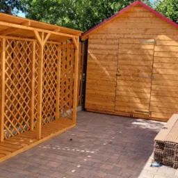 Drewniana altana z kratką i domek narzędziowy z czerwonym dachem na kostce brukowej, otoczone zielenią i drewnianym płotem.