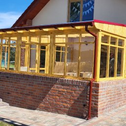 INSTAL-WOOD WIESŁAW SZWED - Zadaszony taras z drewnianą konstrukcją i ceglaną podstawą. Widoczne schody wejściowe i otoczenie posesji w słoneczny dzień. Czerwony dach i rynna.