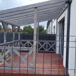 INSTAL-WOOD WIESŁAW SZWED - Zadaszony taras z drewnianą podłogą, balustradą w kształcie krzyży i szarą konstrukcją. Widok przez metalowe ogrodzenie na tle domu z białymi ścianami.