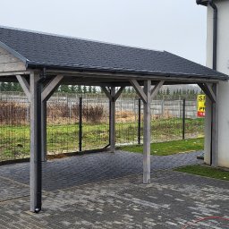 INSTAL-WOOD WIESŁAW SZWED - Drewniany carport z czarnym dachem z gontu bitumicznego, wsparty na czterech słupach, przylegający do białego budynku z oknem. Widoczna kostka brukowa i ogrodzenie.