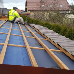 INSTAL-WOOD WIESŁAW SZWED - Montaż brązowej blachodachówki na dachu domu. Widoczny pracownik w żółtej kamizelce i drewniana konstrukcja dachu z niebieską membraną. Częściowo ułożone dachówki.