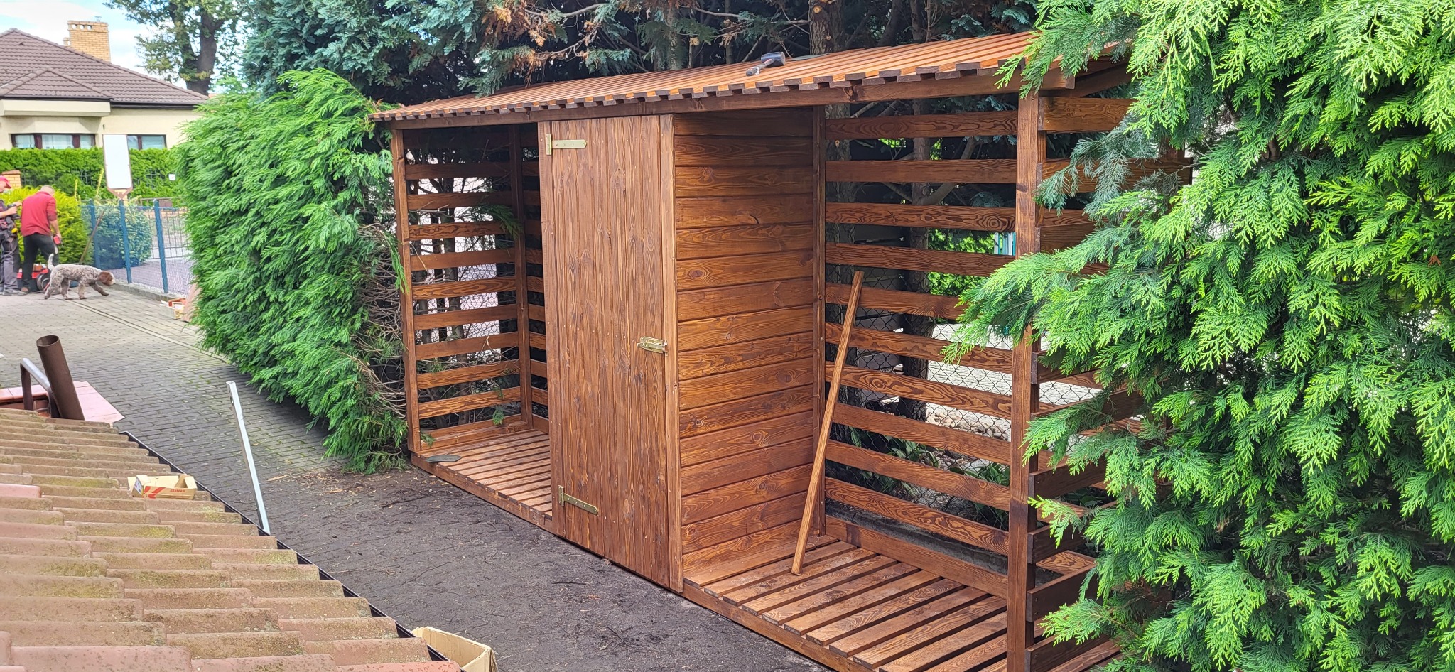 Drewniana, brązowa wiata ogrodowa z drzwiami, z ażurowymi ścianami, stojąca na zewnątrz obok zielonych krzewów i drzew. Widok z góry, z dachu budynku.