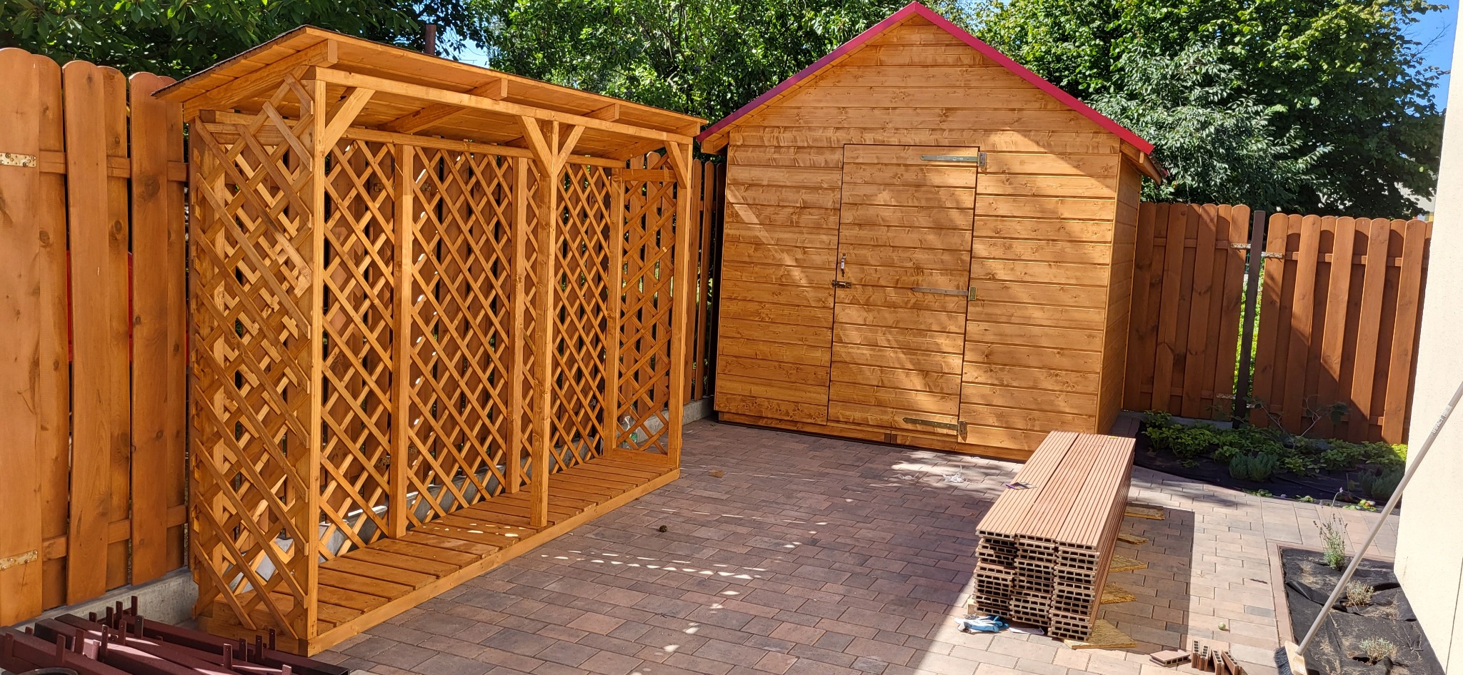 Drewniana altana z kratką i domek narzędziowy z czerwonym dachem na kostce brukowej, otoczone zielenią i drewnianym płotem.