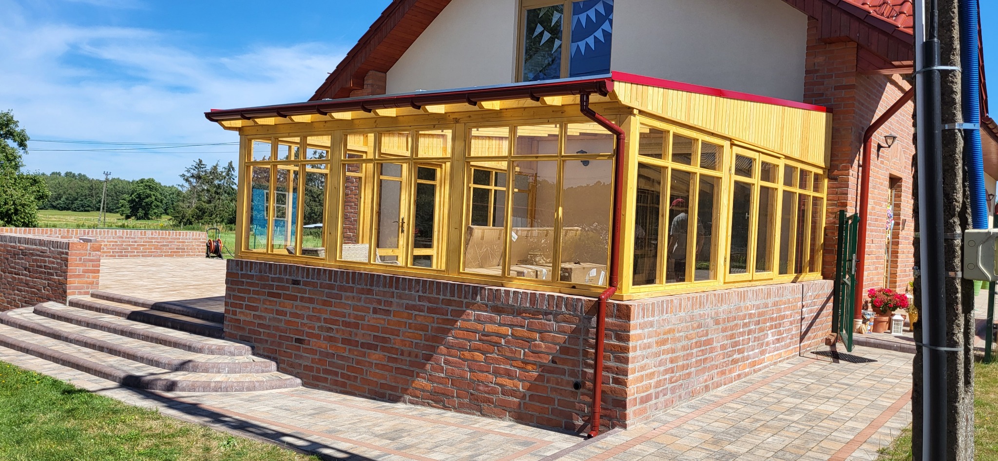 Zadaszony taras z drewnianą konstrukcją i ceglaną podstawą. Widoczne schody wejściowe i otoczenie posesji w słoneczny dzień. Czerwony dach i rynna.