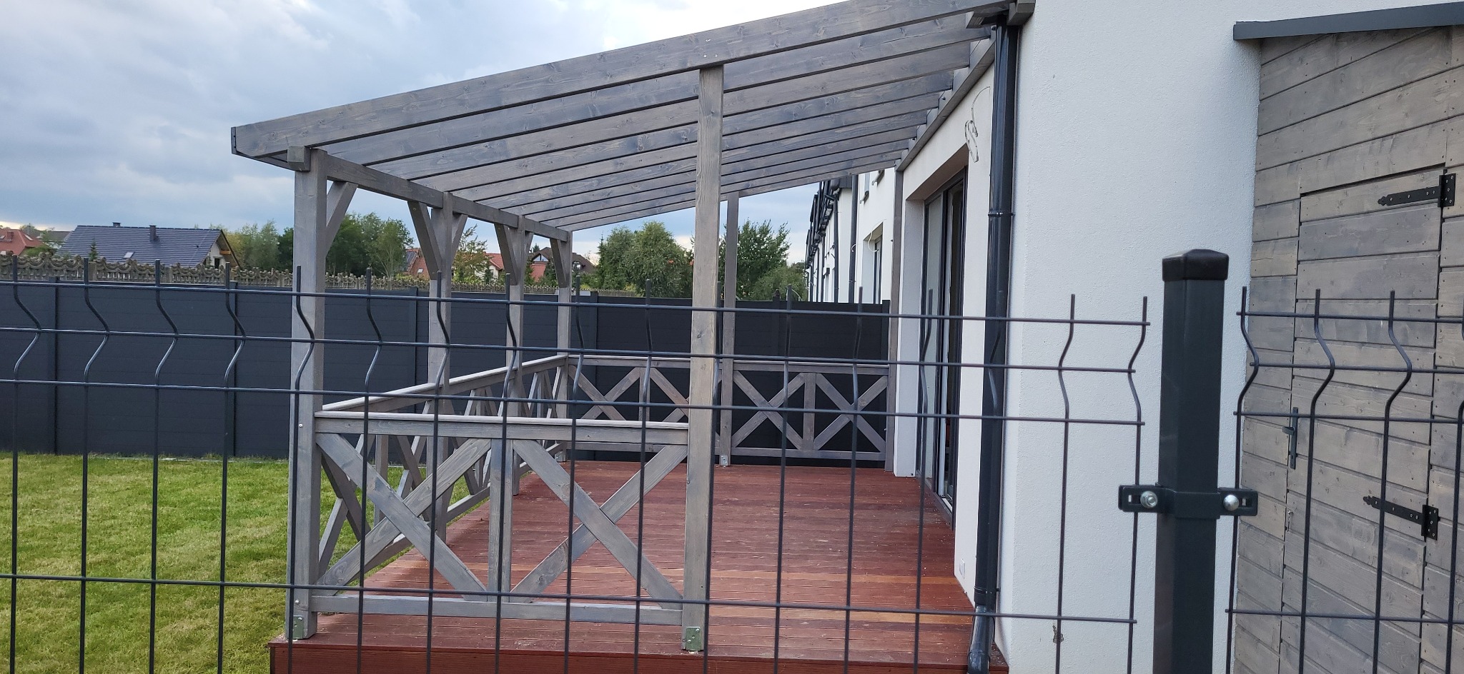 Zadaszony taras z drewnianą podłogą, balustradą w kształcie krzyży i szarą konstrukcją. Widok przez metalowe ogrodzenie na tle domu z białymi ścianami.