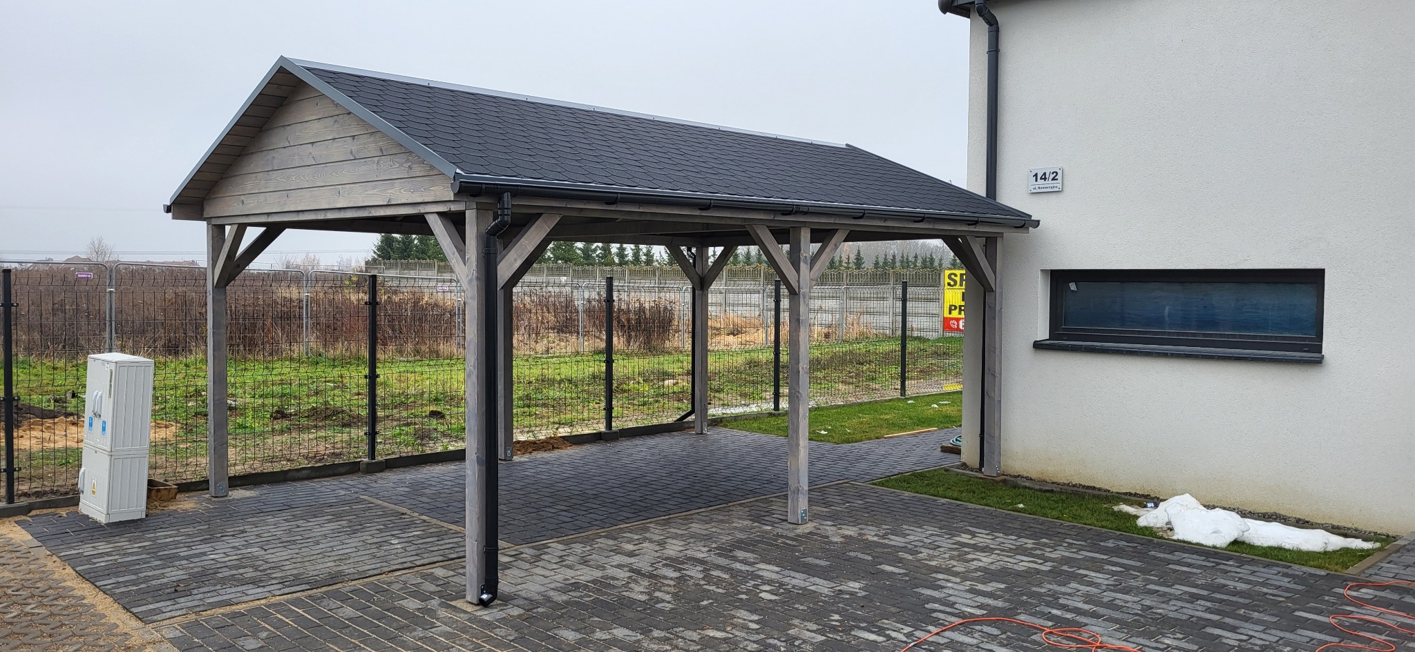 Drewniany carport z czarnym dachem z gontu bitumicznego, wsparty na czterech słupach, przylegający do białego budynku z oknem. Widoczna kostka brukowa i ogrodzenie.