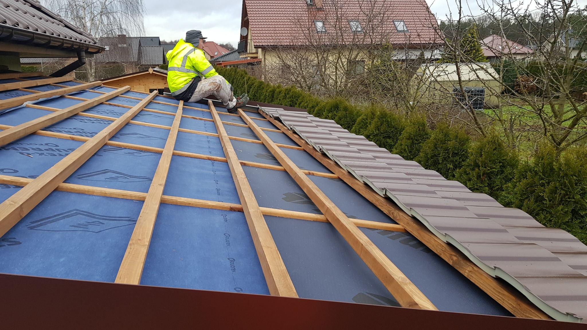 Montaż brązowej blachodachówki na dachu domu. Widoczny pracownik w żółtej kamizelce i drewniana konstrukcja dachu z niebieską membraną. Częściowo ułożone dachówki.