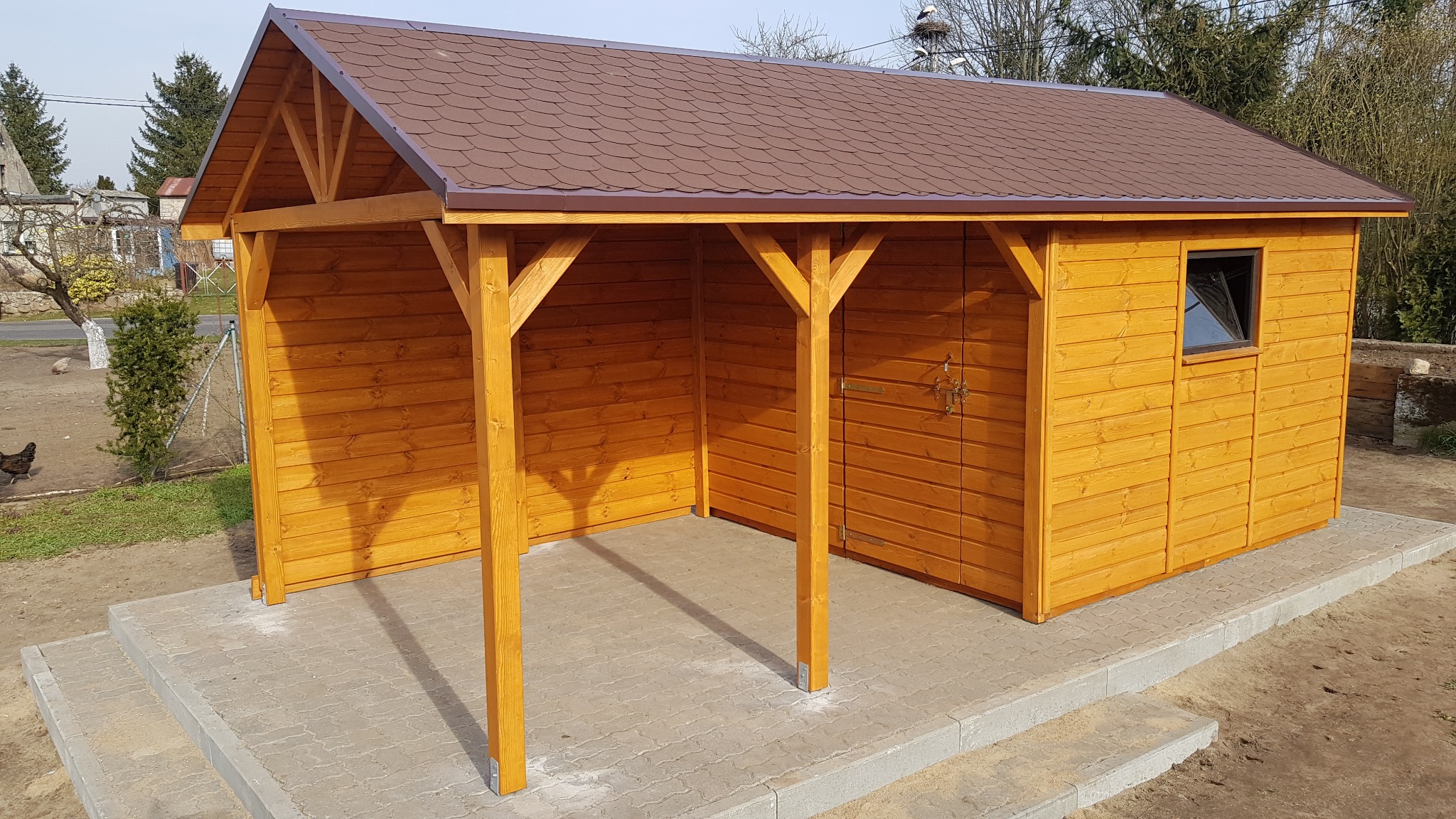 Drewniana altana z brązowym dachem, widoczna z zewnątrz. Konstrukcja z jasnego drewna, zadaszona część i zamknięta z oknem. Podłoże wyłożone kostką brukową.