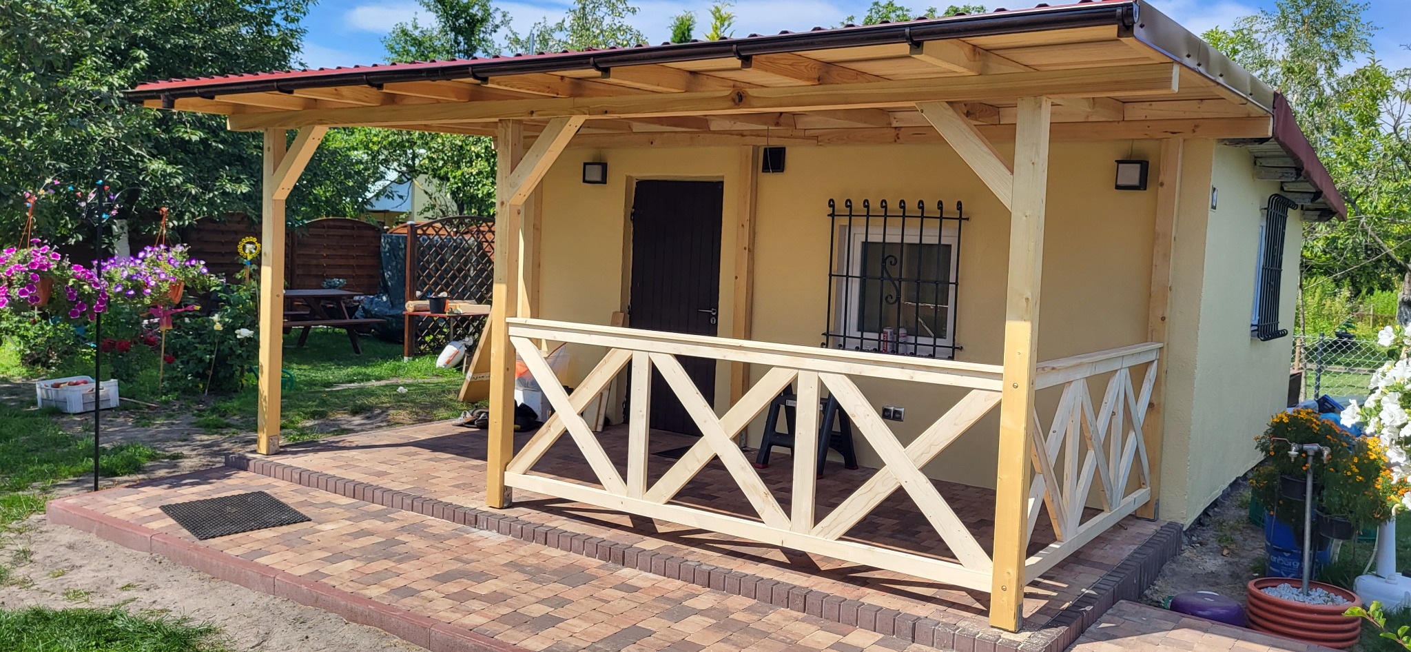 Drewniana altana z beżową elewacją i dekoracyjną balustradą w kształcie litery X, otoczona zielenią ogrodu i kolorowymi kwiatami w donicach.