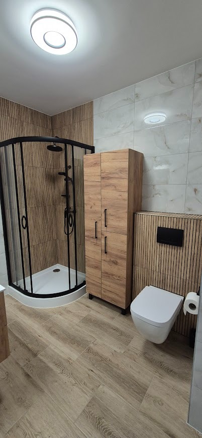 Nowoczesna łazienka po remoncie: kabina prysznicowa z czarnymi profilami, drewnopodobne panele ścienne, szafka i toaleta. Oświetlenie sufitowe.