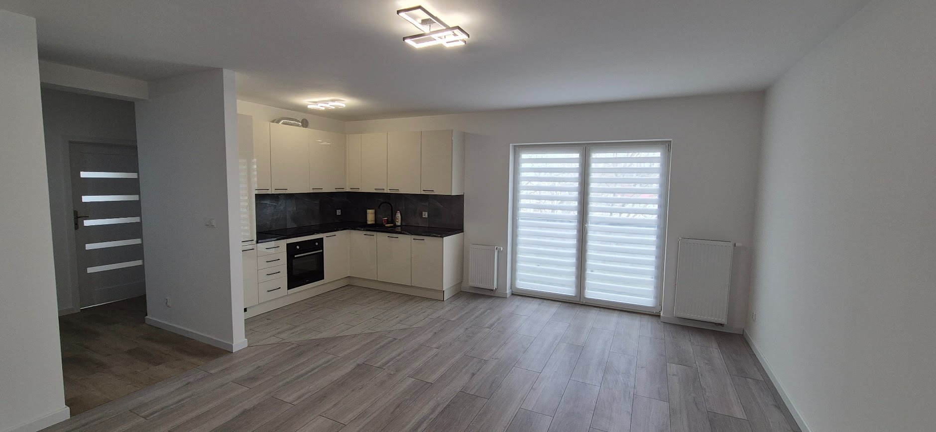 Jasna kuchnia z czarnym blatem i płytkami, widok na okno z roletami i grzejnik. Podłoga w odcieniach szarości imitująca drewno. Nowoczesne oświetlenie sufitowe.