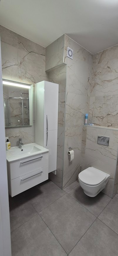 Nowoczesna łazienka z marmurowymi płytkami, białą szafką z umywalką, wiszącą toaletą i szarą podłogą. Minimalistyczny design z akcentami w postaci srebrnych uchwytów.