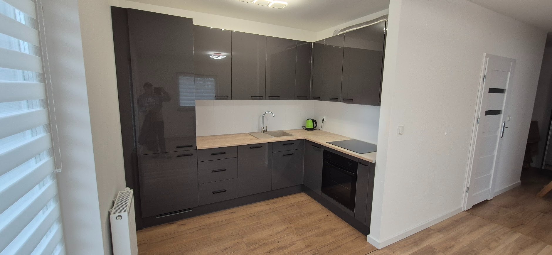 Nowoczesna, szara kuchnia z drewnianym blatem i błyszczącymi frontami szafek. Widoczny zlew, płyta grzewcza i biała ściana z oknem. W tle drzwi.