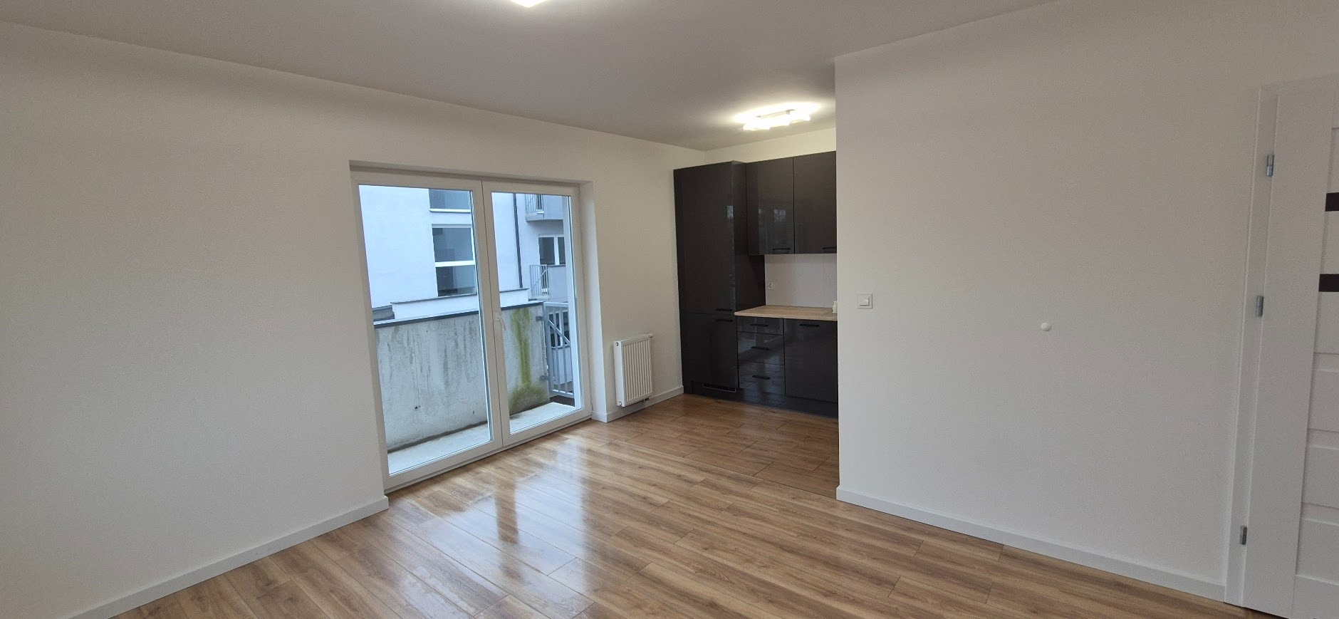 Jasne wnętrze po remoncie: drewnopodobna podłoga, białe ściany, czarna zabudowa kuchenna, okno balkonowe. Minimalistyczny design, świeżo po odnowieniu.