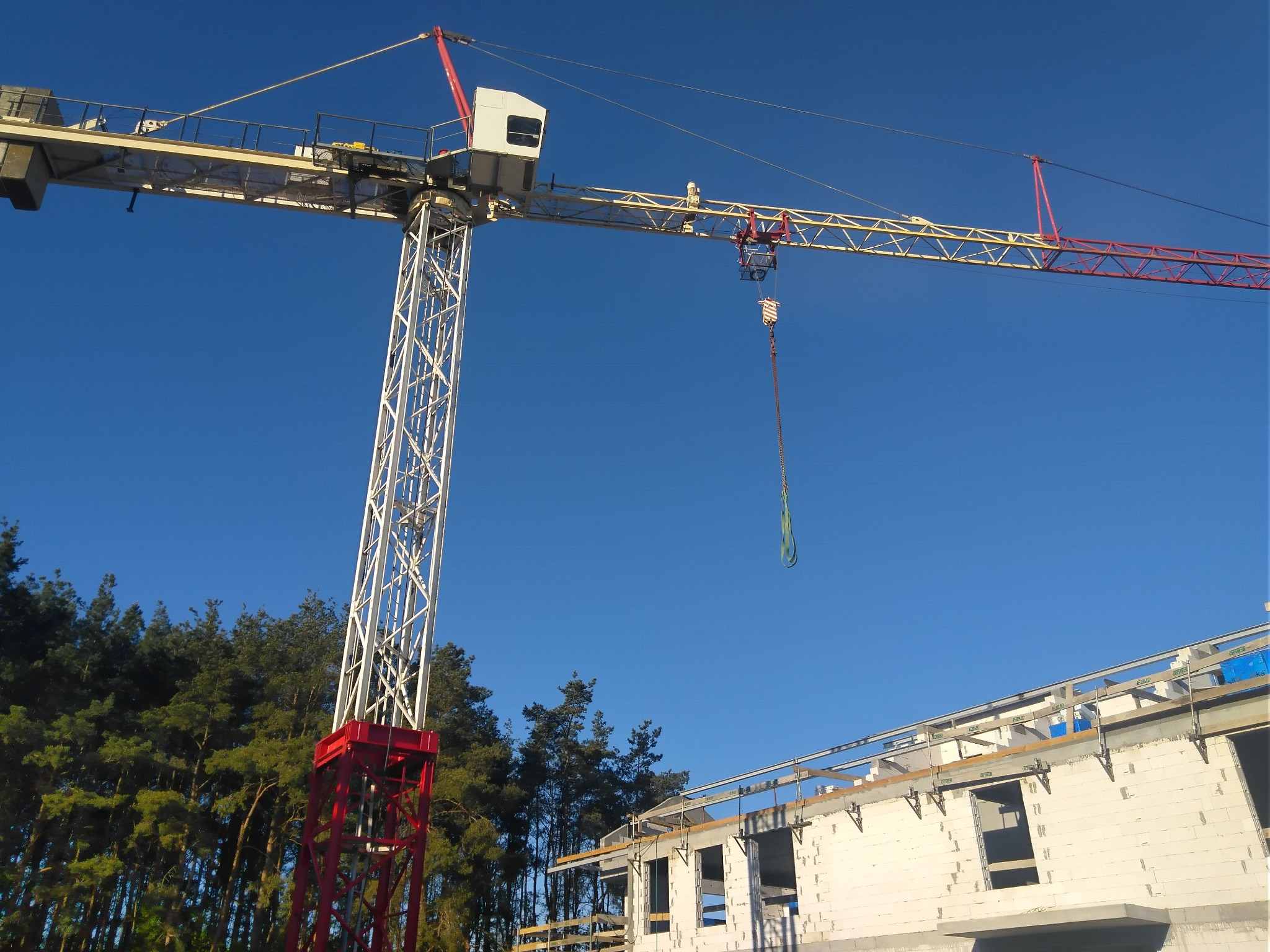 Żuraw wieżowy na tle błękitnego nieba i częściowo wzniesionego budynku z białych bloczków. W tle widoczny las iglasty.