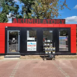 Czerwono-czarny pawilon z napisem 'AKUMULATORY' na dachu, prezentujący ofertę akumulatorów w oknach i na stojaku przed wejściem. Słoneczny dzień.