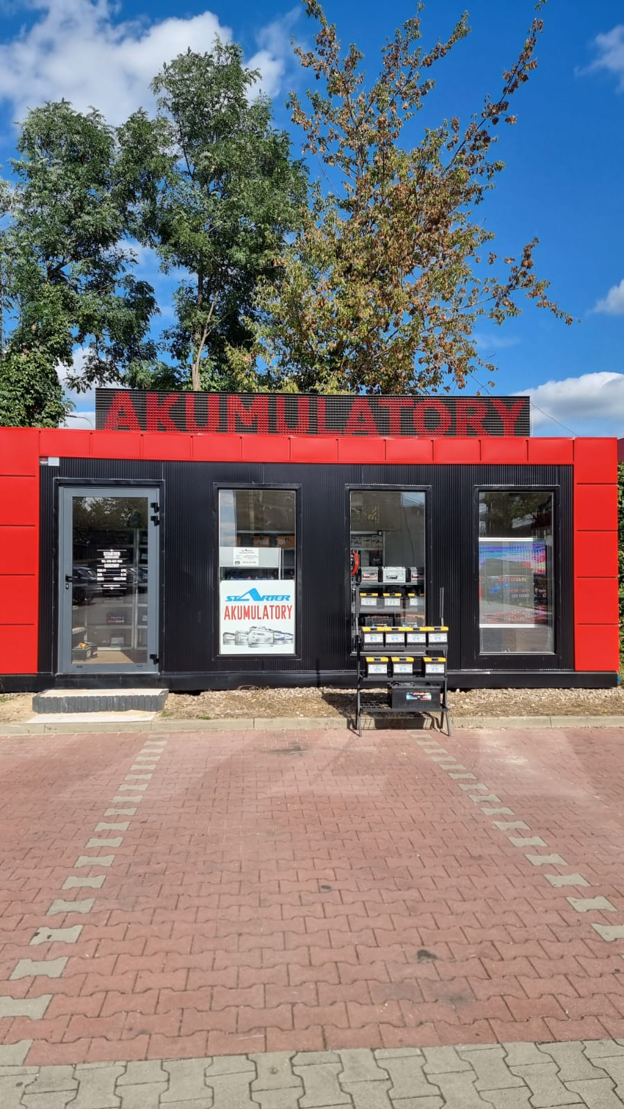 Czerwono-czarny pawilon z napisem 'AKUMULATORY' na dachu, prezentujący ofertę akumulatorów w oknach i na stojaku przed wejściem. Słoneczny dzień.