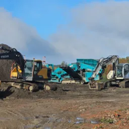 Dwie koparki (Volvo, Terex) i kruszarka na placu budowy z błotnistym podłożem, pod błękitnym niebem z chmurami. Sprzęt ciężki w trakcie pracy.