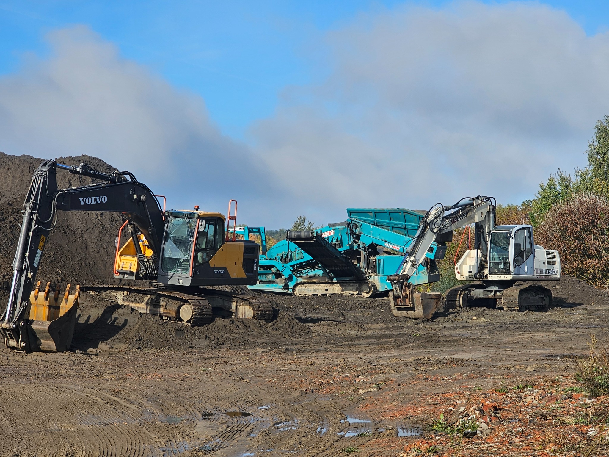 Dwie koparki (Volvo, Terex) i kruszarka na placu budowy z błotnistym podłożem, pod błękitnym niebem z chmurami. Sprzęt ciężki w trakcie pracy.