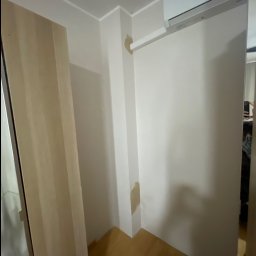 MBworks Mykhailo Balkovyi - Wnętrze pokoju z zamontowaną klimatyzacją na ścianie. Widoczne ślady po naprawach na ścianie, drewniana podłoga i fragment szafy.