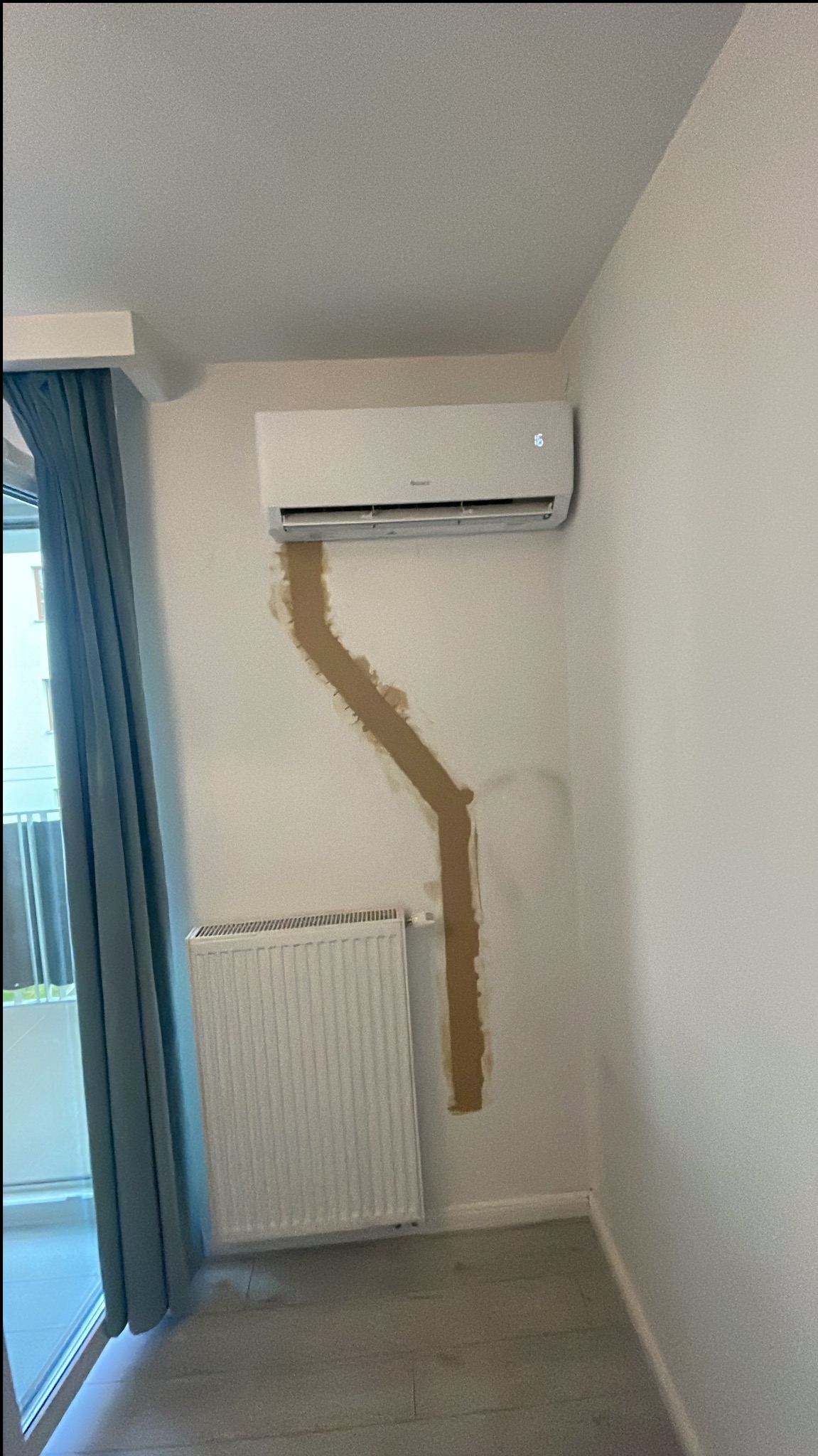 Biały klimatyzator ścienny po montażu, widoczne ślady po instalacji rur na ścianie, obok grzejnik i okno z zasłoną.