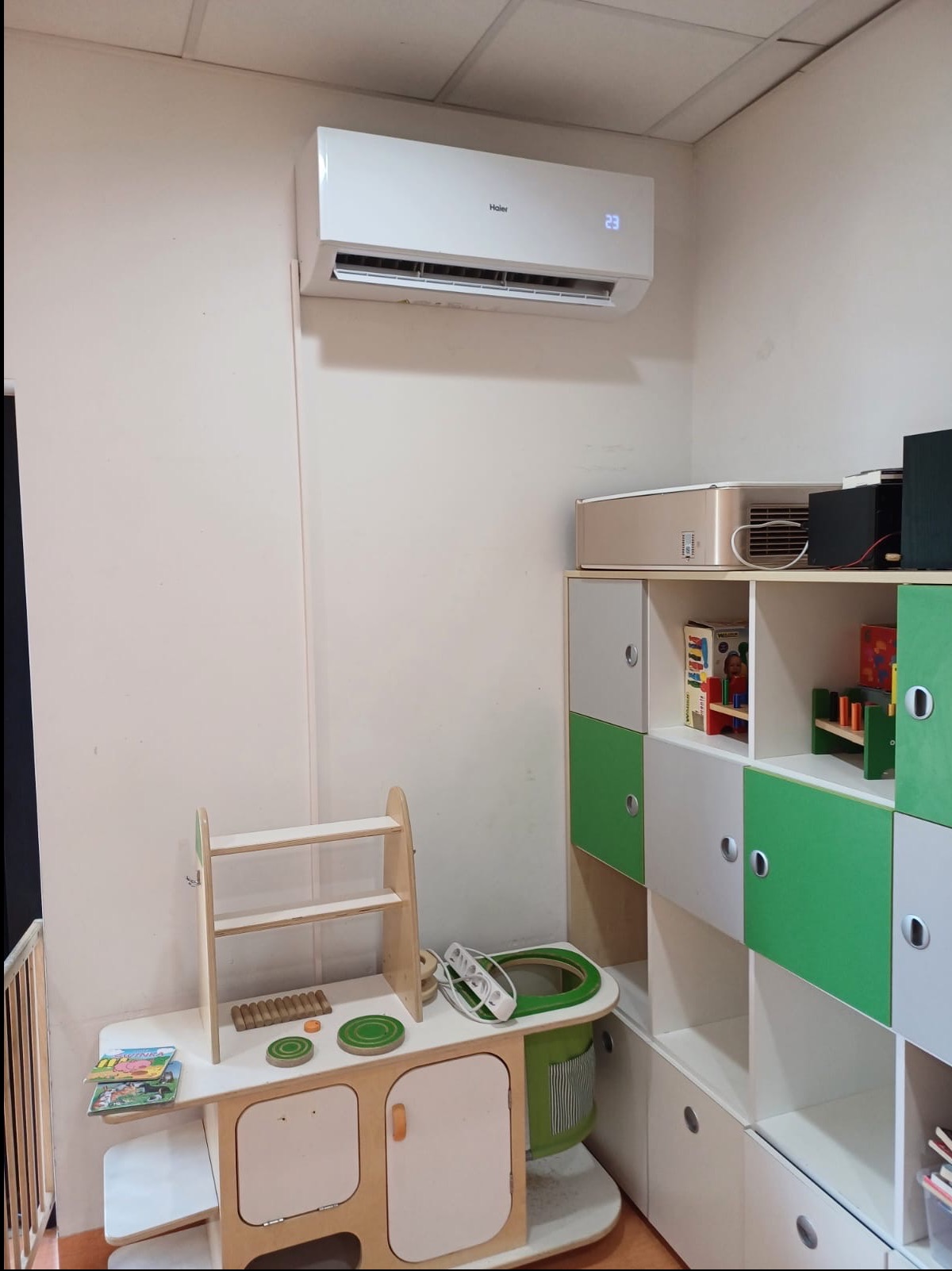 Biały klimatyzator Haier zamontowany na ścianie w pokoju dziecięcym. Widoczny fragment umeblowania dziecięcego i zabawki. Ściana w jasnym odcieniu beżu.