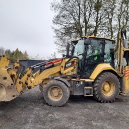 Firma Usługowa JARKOP Tomasz Jarek - Żółta koparko-ładowarka Caterpillar 428F2 na żwirowym podłożu, z widocznym łyżką i ramieniem kopiącym, na tle drzew i zabudowań. Operator w kabinie.