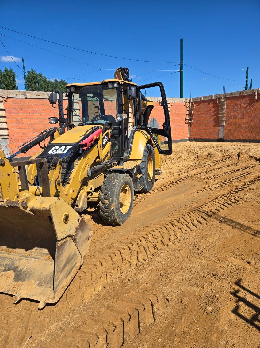 Żółta koparko-ładowarka CAT 428F na placu budowy z fundamentami z czerwonej cegły. Ślady opon na piasku, błękitne niebo w tle. Słoneczny dzień.