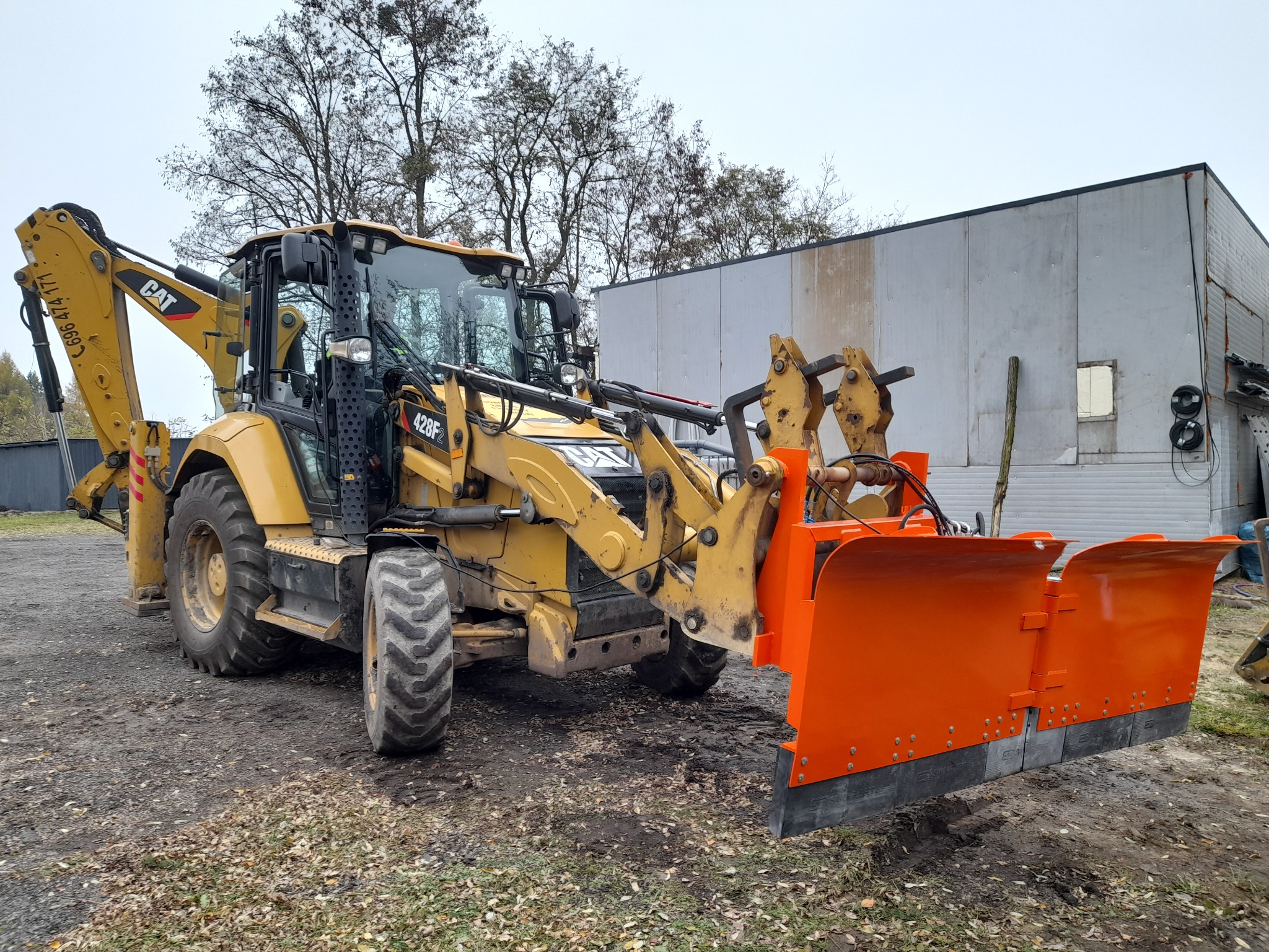 Żółta koparko-ładowarka CAT 428F2 z pomarańczowym pługiem do odśnieżania na błotnistym terenie, z budynkiem w tle. Widoczne detale konstrukcyjne i opony.