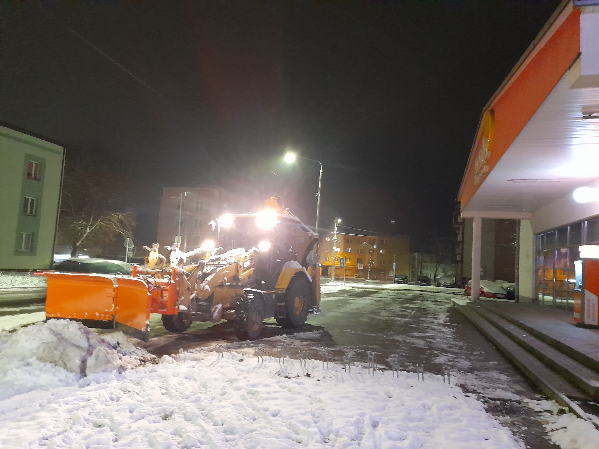 Żółty traktor z pomarańczowym pługiem odśnieża drogę w nocy, oświetlony latarnią. W tle budynki i stacja benzynowa.