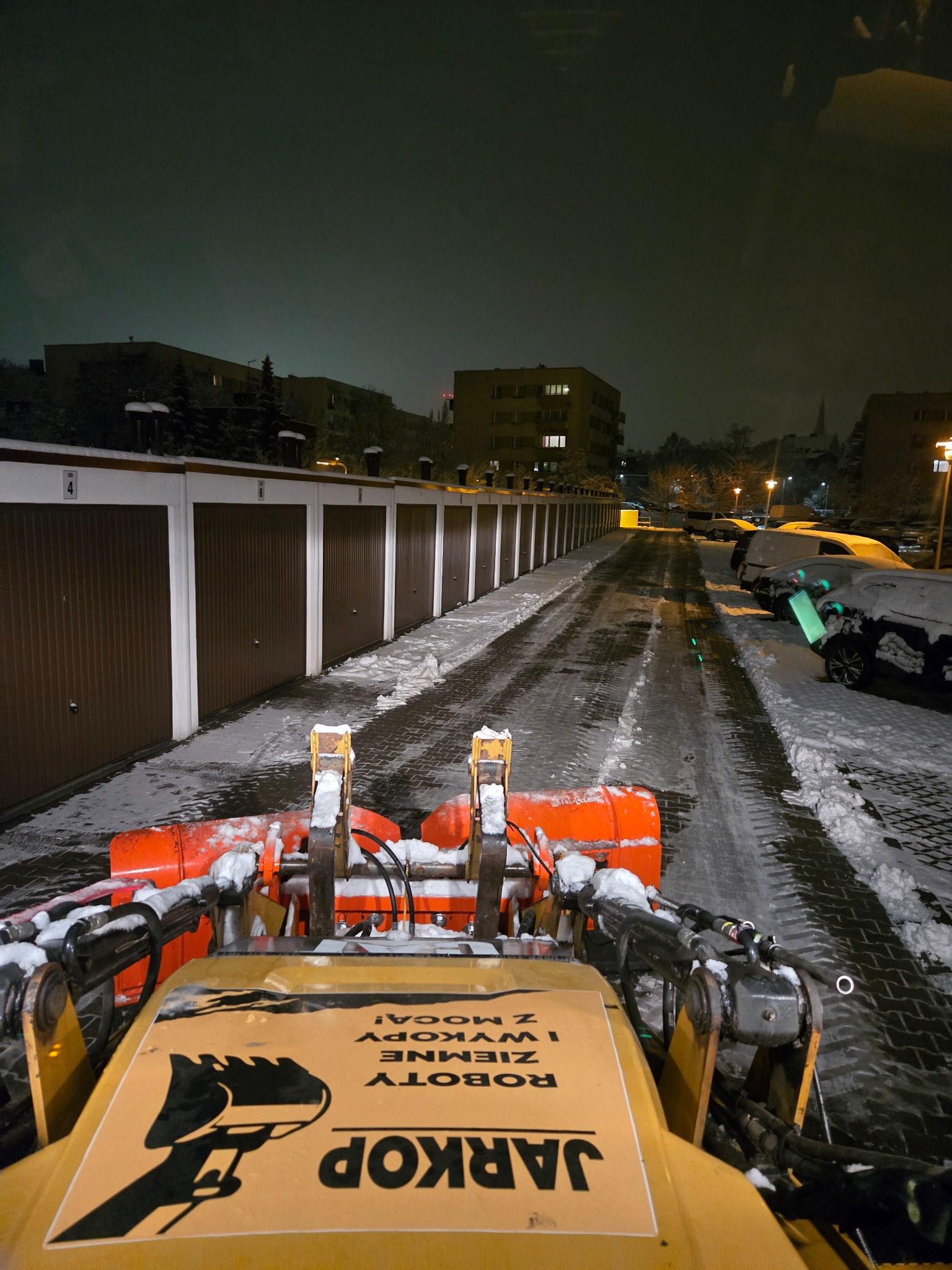 Odśnieżarka z pomarańczowym pługiem na tle zaśnieżonej drogi między garażami, nocne oświetlenie, widok z perspektywy operatora, logo firmy na maszynie.