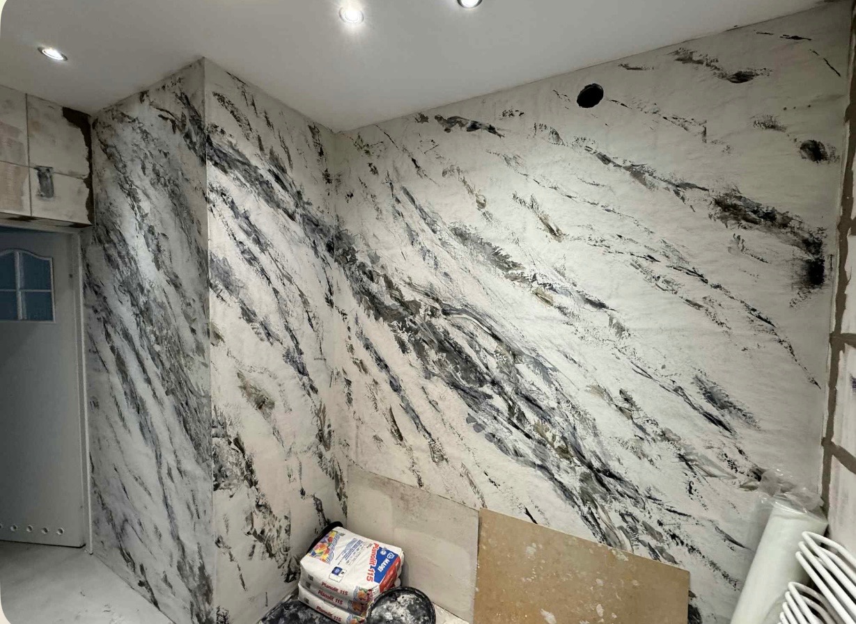 Wykończona ściana w pomieszczeniu z efektem dekoracyjnym imitującym marmur, widoczne materiały budowlane w rogu, białe światło.