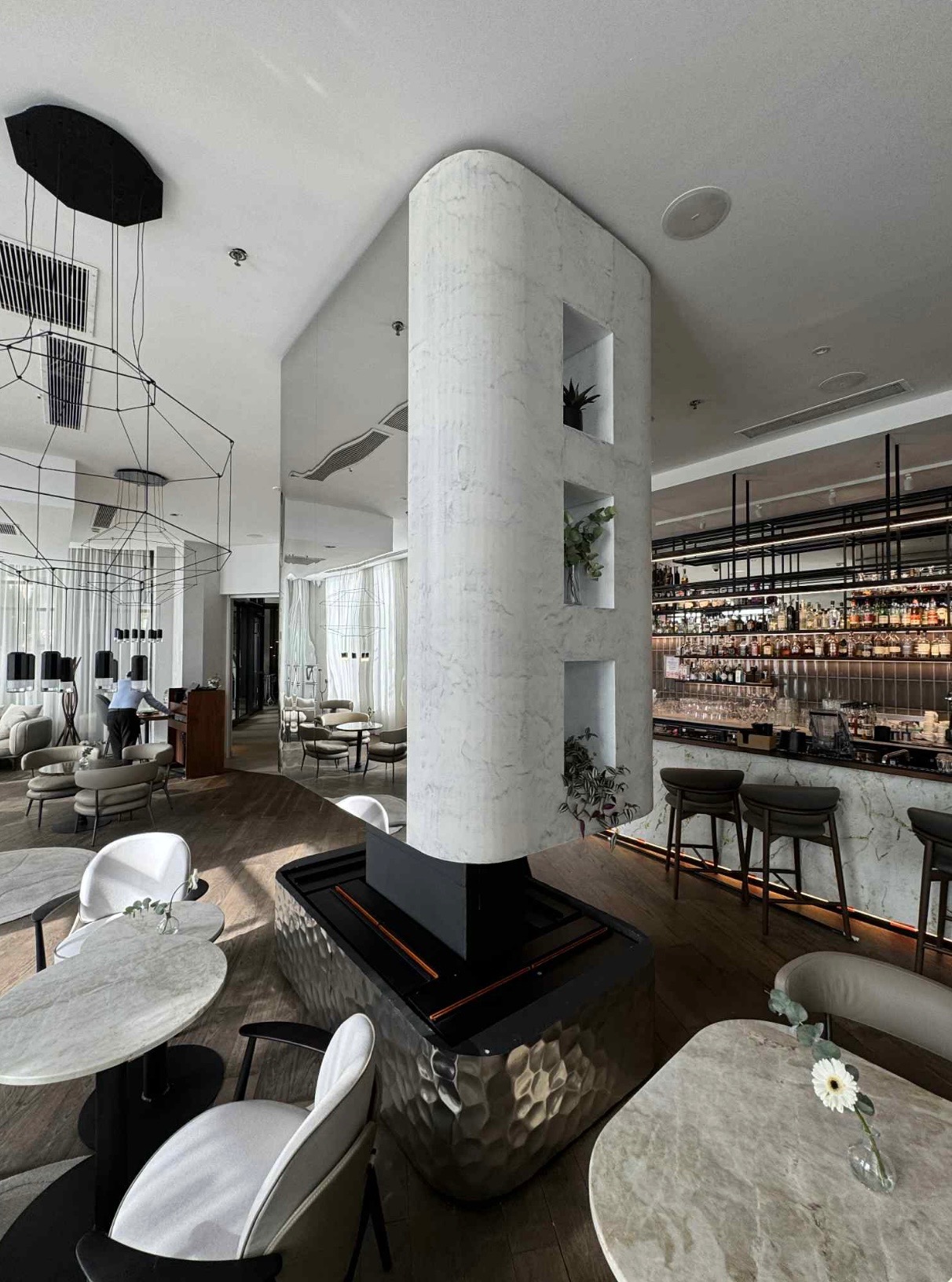 Eleganckie wnętrze baru z marmurową kolumną, roślinami i barkiem w tle. Nowoczesne oświetlenie i drewniana podłoga dodają szyku.